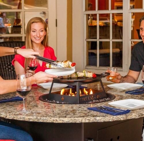 Group of people enjoying a meal around a table with a gas fire pit built-in for cooking.