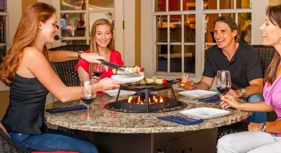 Group of people enjoying a meal around a table with a gas fire pit built-in for cooking.