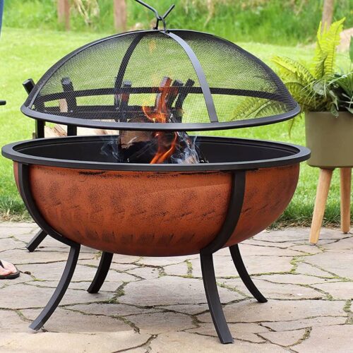 A person stands next to a rust-toned fire pit bowl with a protective mesh screen, burning flames visible, set on a garden patio.