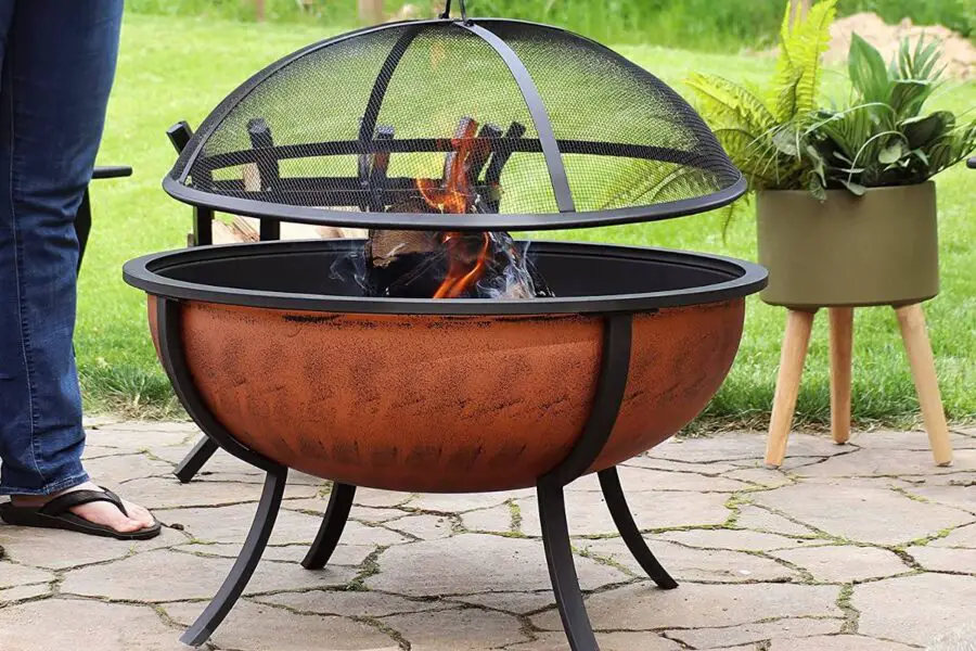 A person stands next to a rust-toned fire pit bowl with a protective mesh screen, burning flames visible, set on a garden patio.