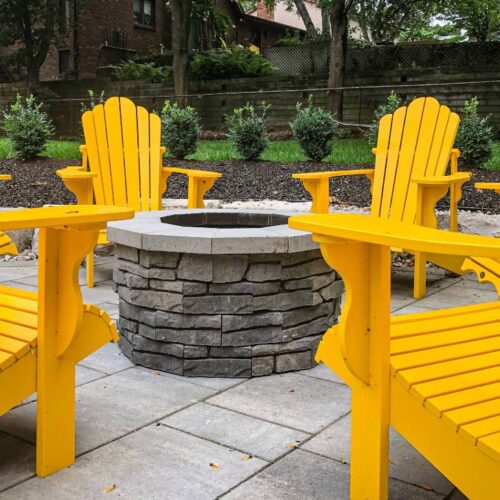 Four yellow adirondack chairs arranged at a safe distance around a circular stone fire pit, complying with safety regulations, in a backyard garden setting.