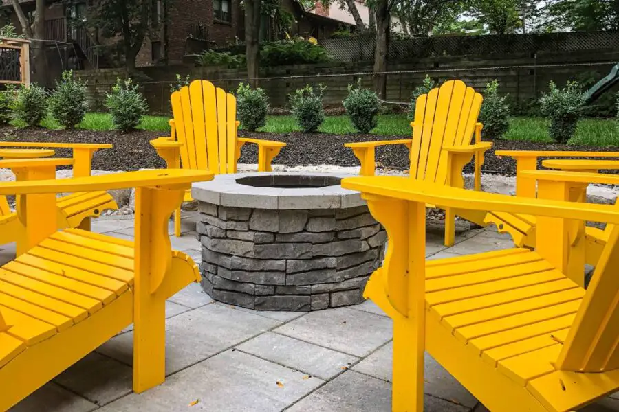 Four yellow adirondack chairs arranged at a safe distance around a circular stone fire pit, complying with safety regulations, in a backyard garden setting.