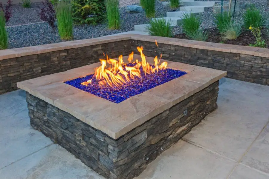 A gas fire pit area with blue glass crystals, surrounded by stone seating in an outdoor setting.