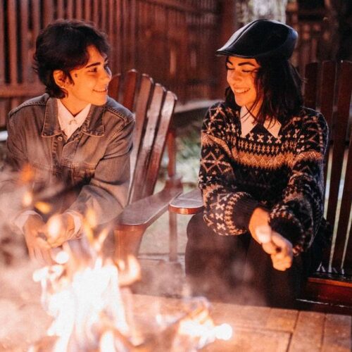 Two individuals enjoying a conversation by a fire pit, surrounded by a fence, at night.