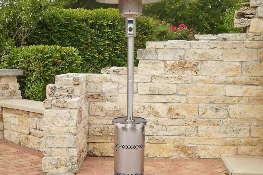 Patio heater standing on a brick pavement near a stone wall with plants, ready for use indoors.