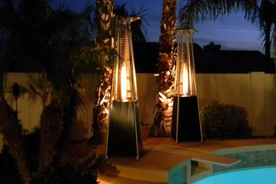 Twin pyramid patio heaters illuminate a poolside area at twilight, casting a warm glow on nearby palm trees.