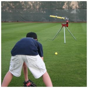 A baseball player crouches in preparation to field a ball launched by a pitching machine on a grassy field.
