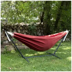 Red hammock on a metal stand in a garden with trees and a woodpile in the background.