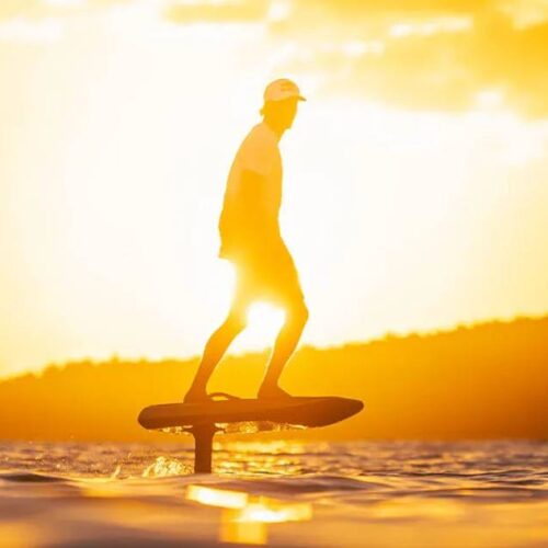 A person using an eFoil board glides across the water at sunset, with the sun casting a golden glow and silhouetting the hills in the background.