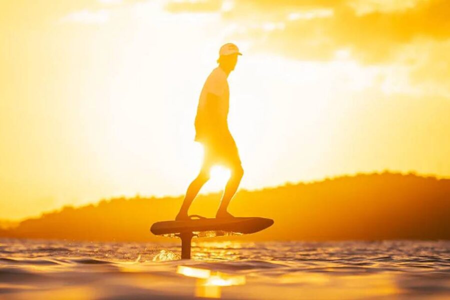A person using an eFoil board glides across the water at sunset, with the sun casting a golden glow and silhouetting the hills in the background.