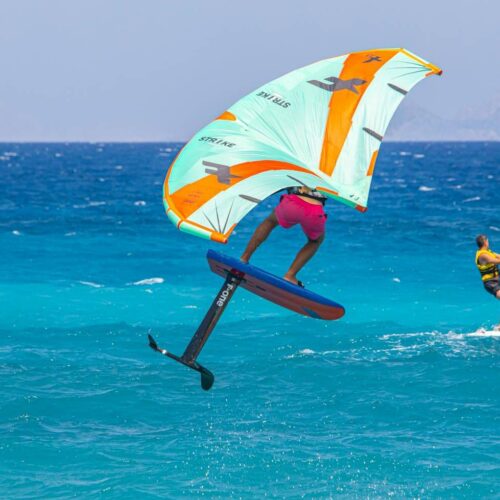 Two people windsurfing in a blue sea, one on a hydrofoil board airborne above the water following a wing foiling guide, and another traditionally surfing nearby.