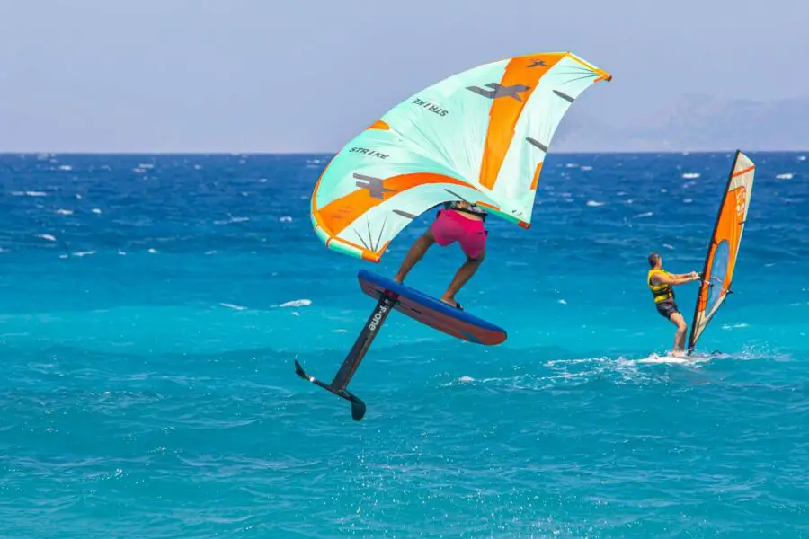 Two people windsurfing in a blue sea, one on a hydrofoil board airborne above the water following a wing foiling guide, and another traditionally surfing nearby.