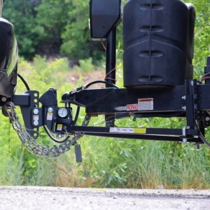 Close-up of a black trailer hitch on a travel trailer, featuring a Weigh Safe Steel Weight Distribution Adjustable Drop Hitch, safety chains, and a propane tank, with greenery in the background.