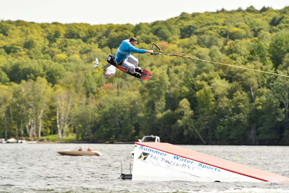 A person in a blue shirt and white helmet performs a jump on a Sky Ski RS Aero 41 Sit-Down Hydrofoil over water, connected by a tow rope, with a ramp and trees in the background.