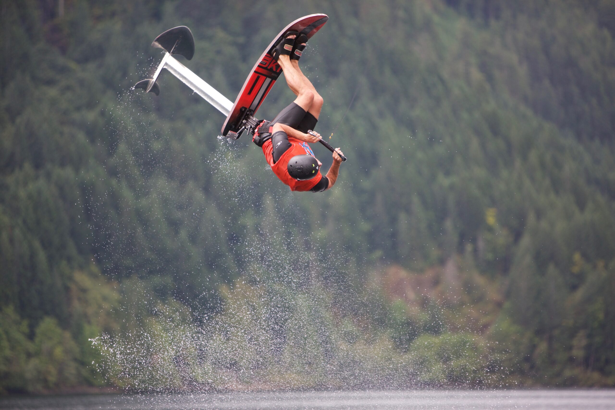 A person performs an aerial flip on a Sky Ski RS Aero 41 Sit-Down Hydrofoil over a body of water with a forested background.