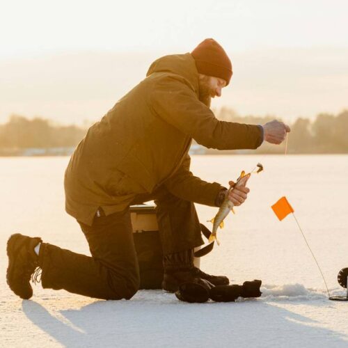 A person, dressed in warm clothing and a beanie, kneels on a frozen lake in Colorado to ice fish, holding a fishing rod near a hole with an orange flag—one of the top picks for winter activities.