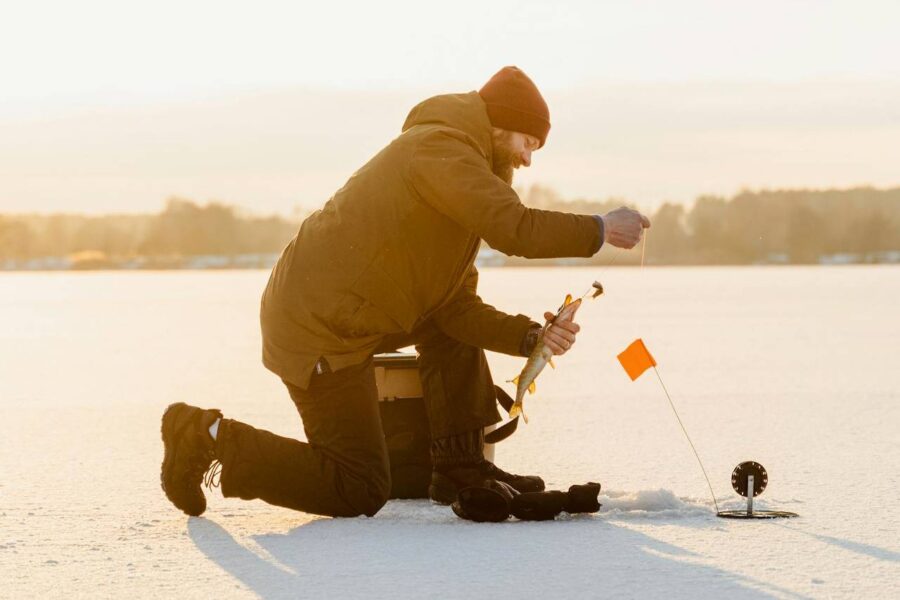 A person, dressed in warm clothing and a beanie, kneels on a frozen lake in Colorado to ice fish, holding a fishing rod near a hole with an orange flag—one of the top picks for winter activities.