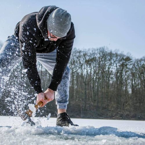 A person in a hoodie and pants is chiseling through the ice on an Illinois lake, surrounded by snow and trees, preparing for a day of ice fishing.