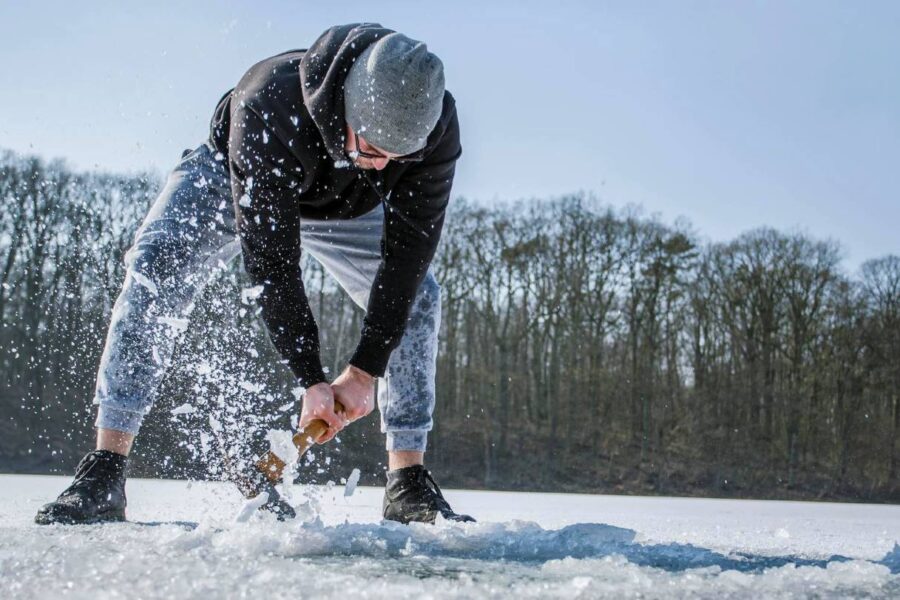 A person in a hoodie and pants is chiseling through the ice on an Illinois lake, surrounded by snow and trees, preparing for a day of ice fishing.