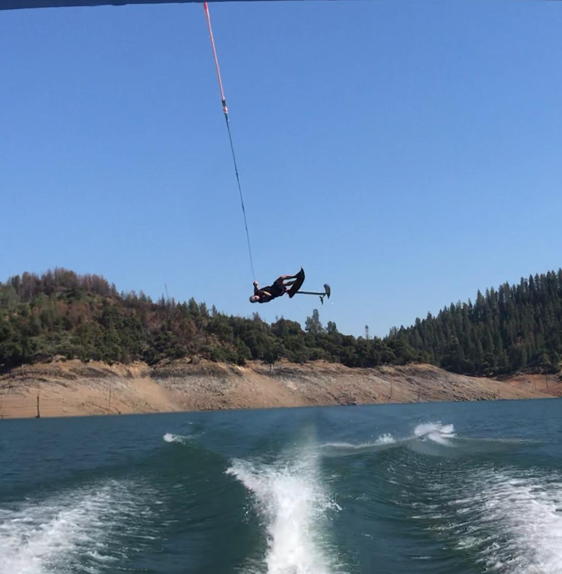 A person is airborne while parasailing over a body of water, connected to a speedboat by a tow rope. Forested hills and a rocky shoreline are visible in the background under a clear sky, with a Sky Ski RS Aero 41 Sit-Down Hydrofoil gliding gracefully below.