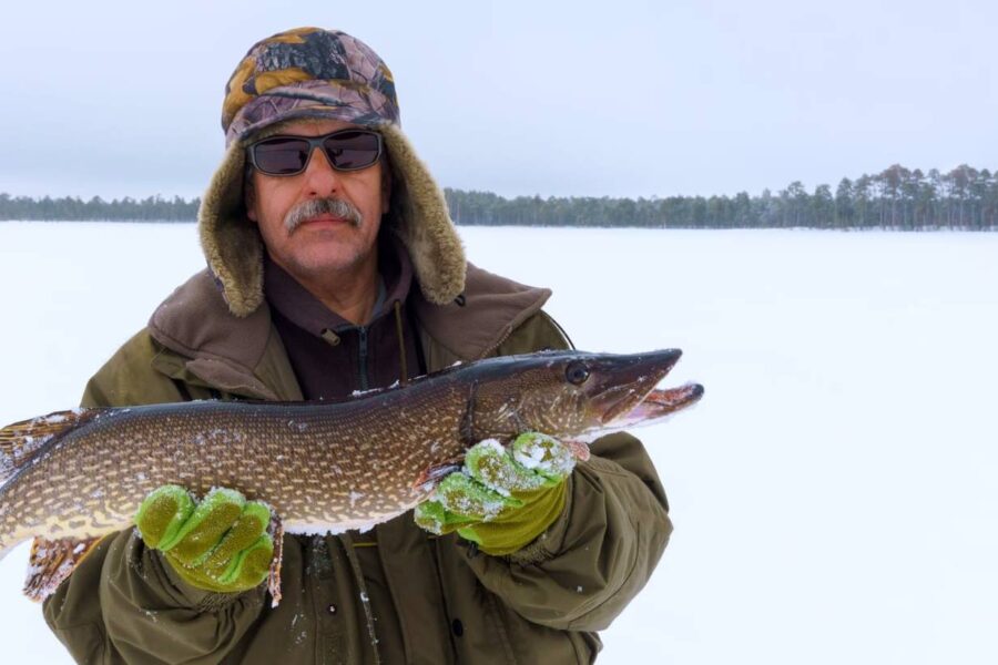 A person bundled in winter gear and sunglasses proudly holds a large fish, standing on a snowy landscape popular for ice fishing in North Dakota.