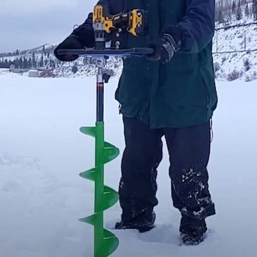 A person operating a green ice fishing auger with a drill attachment on a snowy frozen lake, surrounded by gear and a sled from one of the best ice fishing brands.