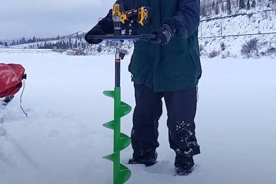 A person operating a green ice fishing auger with a drill attachment on a snowy frozen lake, surrounded by gear and a sled from one of the best ice fishing brands.