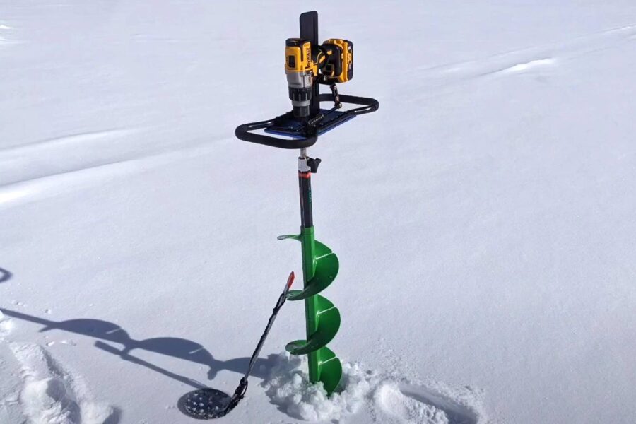 A green ice fishing auger with a yellow and black drill attachment stands upright in the snow, alongside a slotted scoop. This efficient tool can be paired with battery-powered drills for a seamless ice cutting experience.