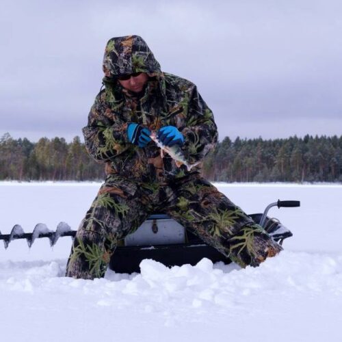 A person in camouflage winter gear stands on a snowy landscape, ice fishing with a drill. A serene lake and the dense forest of Minnesota can be seen in the background.