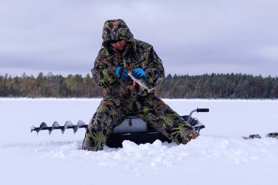 A person in camouflage winter gear stands on a snowy landscape, ice fishing with a drill. A serene lake and the dense forest of Minnesota can be seen in the background.