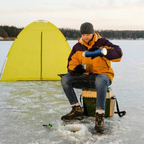 A person sits on a stool on a frozen Wisconsin lake, pouring a drink from a thermos, with a yellow tent, fishing rod, and ice auger nearby, ready for an ice fishing adventure.