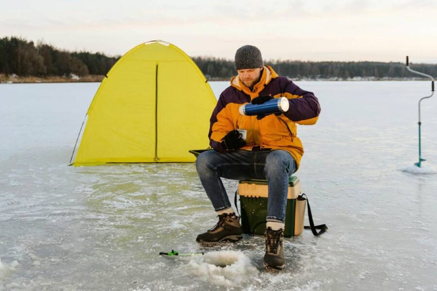 A person sits on a stool on a frozen Wisconsin lake, pouring a drink from a thermos, with a yellow tent, fishing rod, and ice auger nearby, ready for an ice fishing adventure.