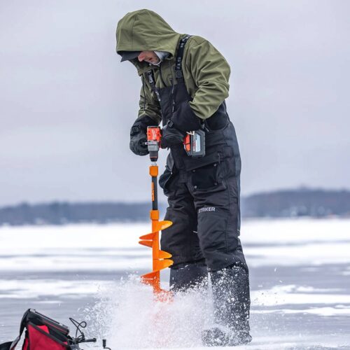 A person in winter gear and ice fishing bibs drills a hole in the ice with an orange auger on a frozen body of water. A red bag lies nearby on the ice.