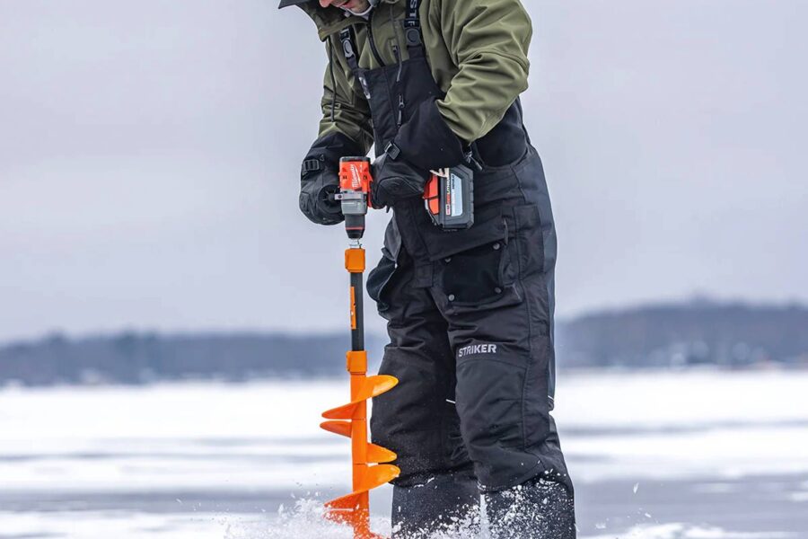 A person in winter gear and ice fishing bibs drills a hole in the ice with an orange auger on a frozen body of water. A red bag lies nearby on the ice.