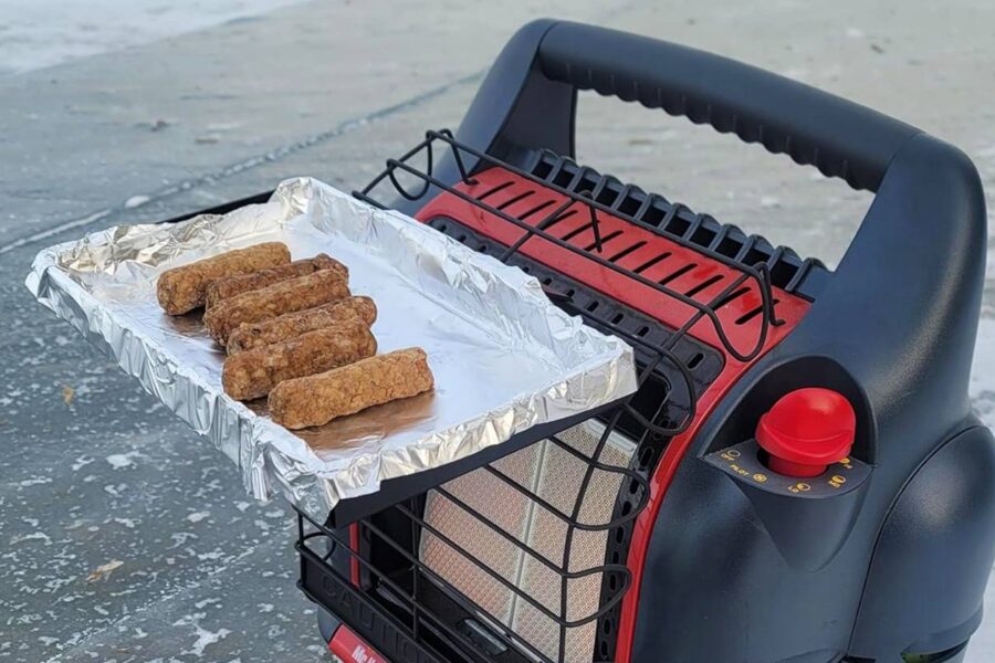 A small portable heater with a wire rack is toasting several sausages on top of a foil tray, perfect for those chilly ice fishing days inside an ice fishing tent.