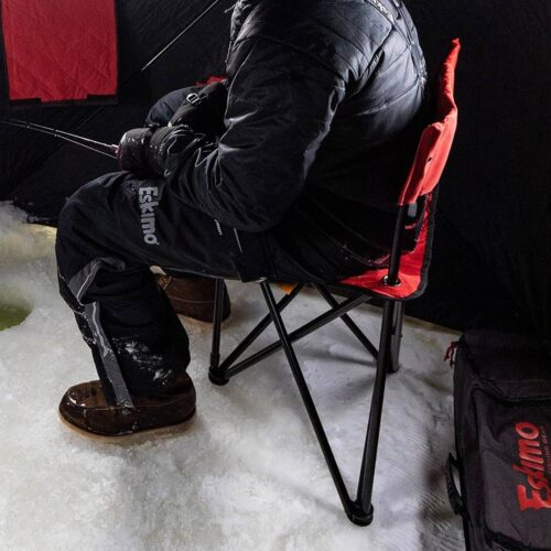 Person in winter clothing ice fishing while seated on one of our top picks for ice fishing chairs—a comfortable red camping chair, with fishing gear and equipment around inside an ice fishing shelter, ensuring frozen comfort.