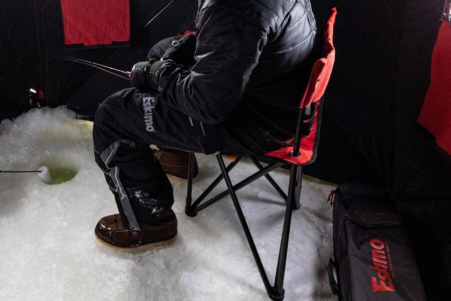 Person in winter clothing ice fishing while seated on one of our top picks for ice fishing chairs—a comfortable red camping chair, with fishing gear and equipment around inside an ice fishing shelter, ensuring frozen comfort.