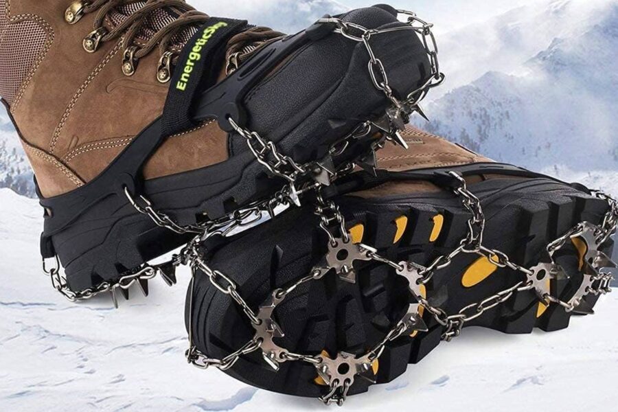 A close-up of boots equipped with metal ice fishing cleats, designed for traction on snowy and icy terrain, with a snowy mountain landscape in the background.