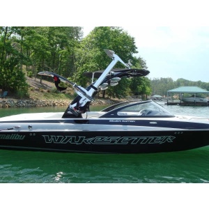 A black and white Malibu Wakesetter boat with a wakeboard tower and a Comptech Marine Pivoting Hydrofoil Rack by Sky Ski is floating on a lake, surrounded by green foliage and other boats in the distance.