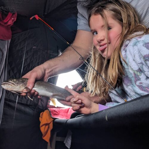 A person shows a fish to a young girl inside a tent. She looks at it with interest, wearing a colorful outfit. Fishing gear and clothing are visible in the background, suggesting they're well-prepared anglers enjoying one of their best ice fishing trips.