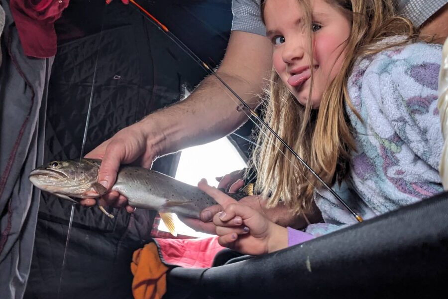 A person shows a fish to a young girl inside a tent. She looks at it with interest, wearing a colorful outfit. Fishing gear and clothing are visible in the background, suggesting they're well-prepared anglers enjoying one of their best ice fishing trips.