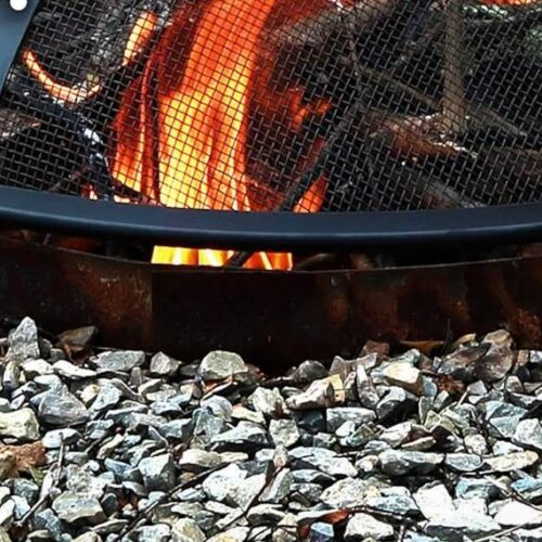 Close-up of a fire pit with burning logs, surrounded by a circular arrangement of gravel stones. The flame is contained by a metal mesh cover—your guide to essential fire pit accessories.