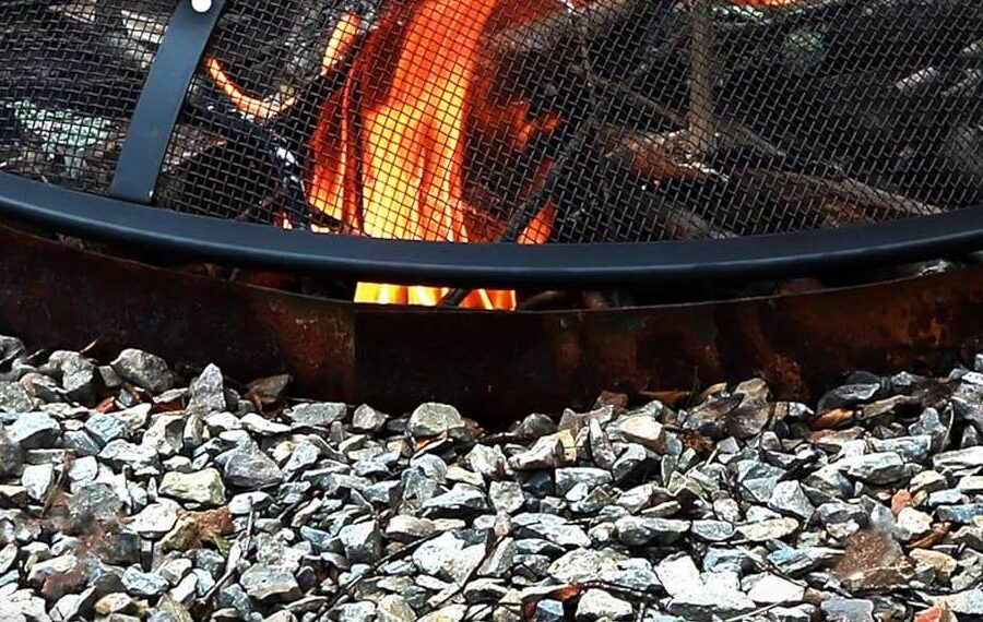 Close-up of a fire pit with burning logs, surrounded by a circular arrangement of gravel stones. The flame is contained by a metal mesh cover—your guide to essential fire pit accessories.