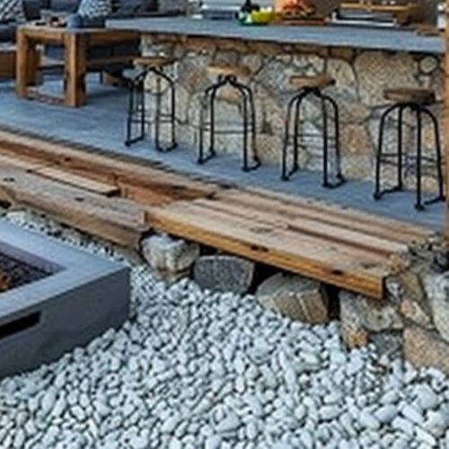 A backyard patio with a stone and wood bar area featuring barstools, a grill, and a sofa. In the foreground, there is a square stone fire pit on a bed of white pebbles with a lit fire, making it essential to ensure regular fire pit maintenance for safety and longevity.