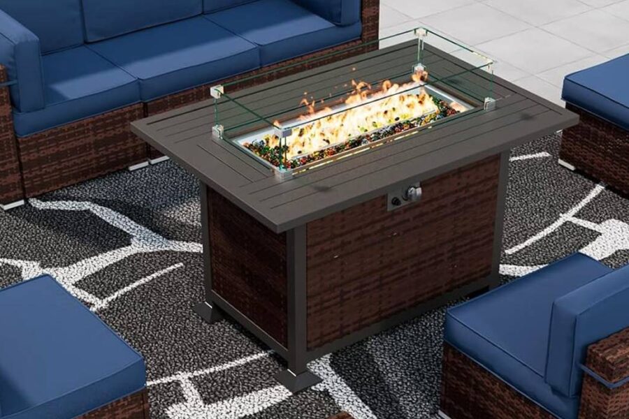 This outdoor patio set, a must-have for outdoor get-togethers, features a wicker fire pit table surrounded by a sofa, two chairs, and an ottoman with blue cushions. All are placed on a patterned rug near the swimming pool—perfect cozy fire pit furniture for any occasion.