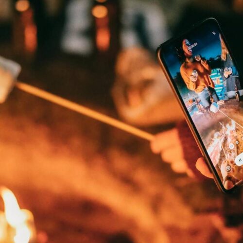 A person holds a smartphone to take a picture of roasting marshmallows over an open fire, mindful of fire pit safety.