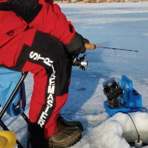 Person in red and black gear ice fishing on a frozen lake, sitting on a folding chair next to a fishing hole with equipment nearby. For the best experience, don't forget your ultimate guide to ice fishing and some of the best ice fishing gloves to keep warm.