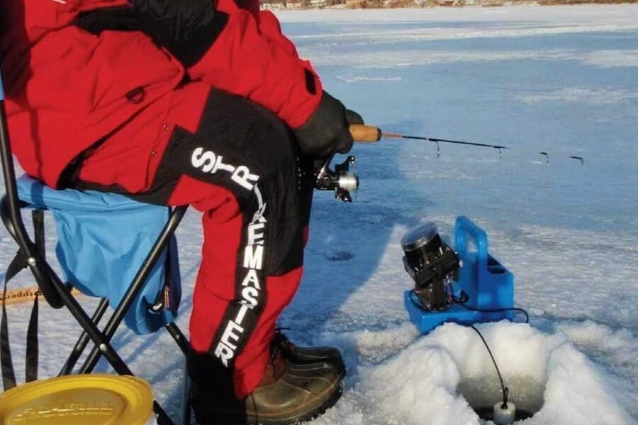 Person in red and black gear ice fishing on a frozen lake, sitting on a folding chair next to a fishing hole with equipment nearby. For the best experience, don't forget your ultimate guide to ice fishing and some of the best ice fishing gloves to keep warm.