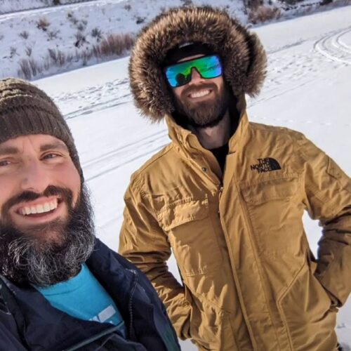 Two bearded men in winter clothing stand outside in a snowy landscape, smiling at the camera. One wears a green knit cap, and the other sports an Angler Coat with a fur-lined hood and reflective sunglasses—clearly among the top picks for best ice fishing coats.