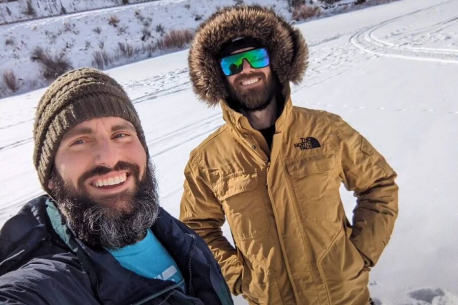 Two bearded men in winter clothing stand outside in a snowy landscape, smiling at the camera. One wears a green knit cap, and the other sports an Angler Coat with a fur-lined hood and reflective sunglasses—clearly among the top picks for best ice fishing coats.